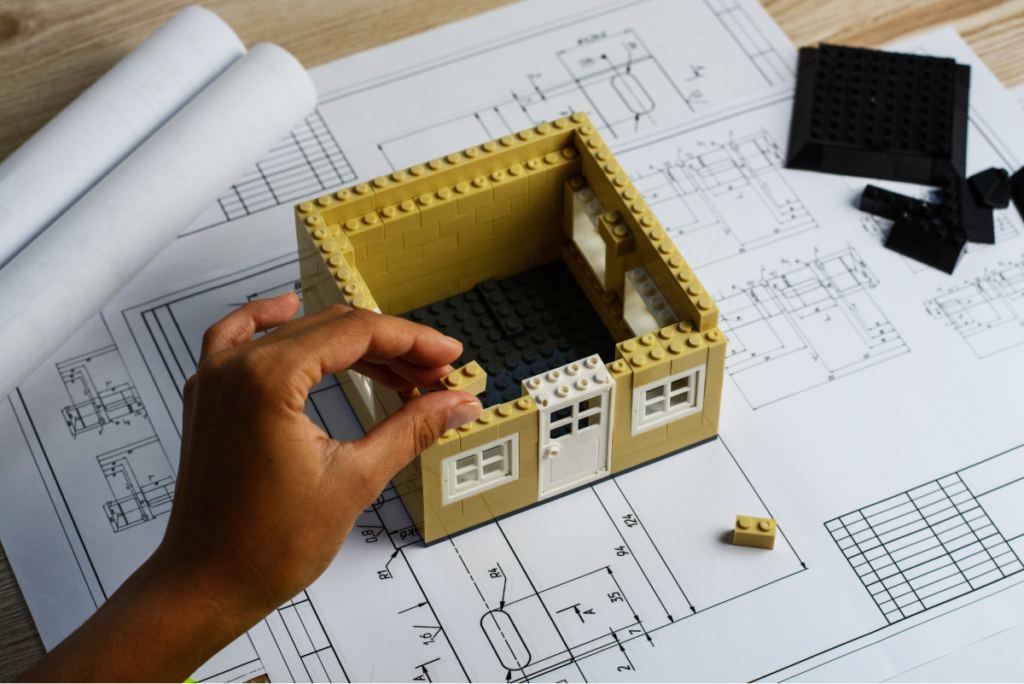 Imagem aproximada na mão de uma pessoa montando uma maquete sobre a planta de uma casa. Arquitetura.