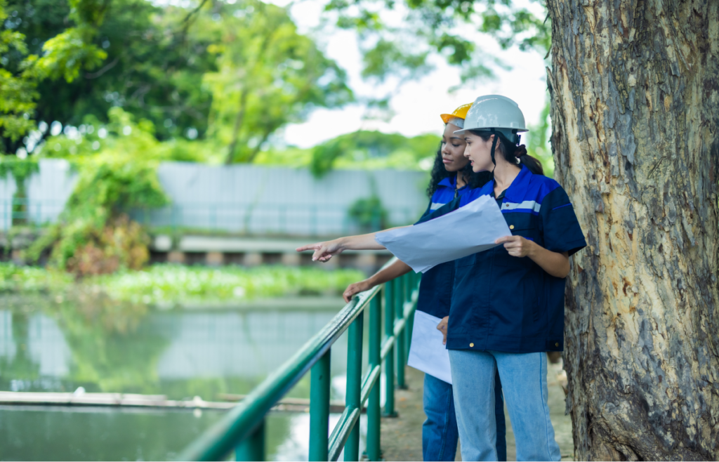 Duas mulheres usando capacete de obra e segurando uma planta conversam e apontam elementos da área. Arquitetura.