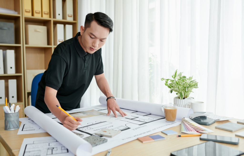 Homem olha para uma planta aberta na mesa e faz anotações com um lápis. Arquitetura.