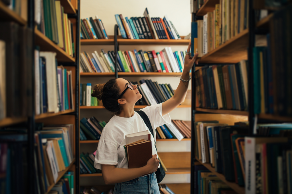 Mulher jovem em biblioteca olhando livros. Redação Enem.
